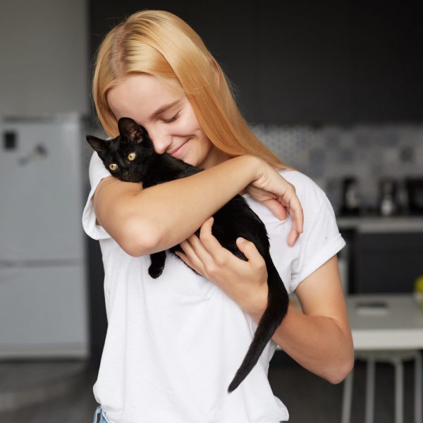 Close up of young blonde girl at the kitchen holds on hands, caresses, stroking, hugs black little cat, cute tenderly smiling, eyes closed, dressed in a domestic white T-shirt, kitchen background